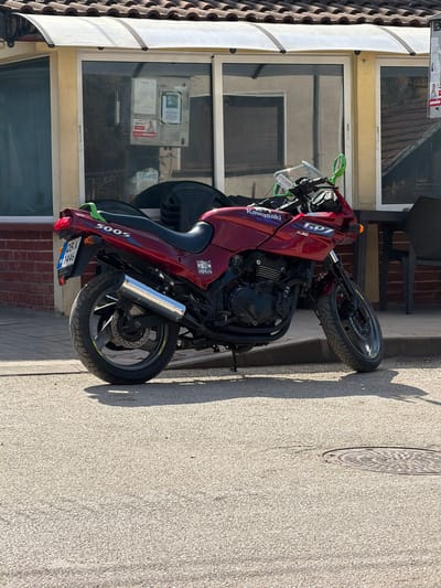 The scene depicts a dark red Kawasaki GPZ 500S motorcycle parked on a light-colored asphalt street in Gradezhnitsa, Bulgaria. The motorcycle is positioned facing the viewer's left, supported by its kickstand, and features green handlebar grips, mirror surrounds, and a rear grab handle. A chrome exhaust pipe is visible on the right side, and the rear license plate reads "0B K 1446". Behind the motorcycle stands a single-story building with a light-colored facade, topped by a terracotta-tiled roof and a clear, arched awning extending over large glass windows. Reflections in the windows show surrounding structures and foliage, and several notices or posters are visible inside or affixed to the windows. Stacked black chairs and a dark table are present to the right of the motorcycle, suggesting an outdoor seating area. The asphalt surface includes a visible circular metal manhole cover in the lower right. The environment is brightly lit by direct sunlight, casting distinct shadows from the motorcycle and the building. No people are present in the image.