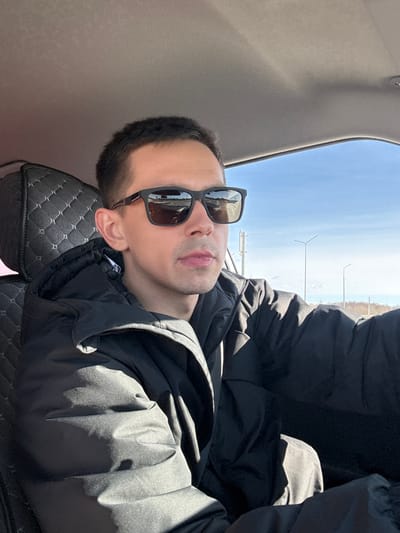 A male individual, appearing to be in his 20s or 30s, is seated in the driver's position of a vehicle in Kukushtan, Russia, looking forward and slightly to his right. He has short, dark hair and is wearing dark-framed rectangular sunglasses, which reflect a segment of a road with visible markings and distant vehicles or structures. He is dressed in a black puffer jacket over a dark undergarment. Strong direct sunlight illuminates his left side, casting defined shadows on his jacket and within the car's interior. The vehicle's interior features a light-colored headliner and a dark, diamond-quilted seat cover on the seat behind him. Through the side window, a clear, bright blue sky is visible above a distant, sparsely vegetated landscape containing several tall utility or light poles. The scene suggests a clear, sunny daytime environment on an open road.