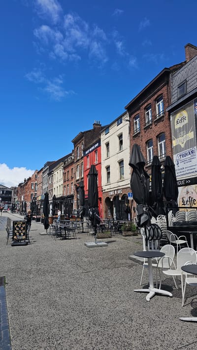 The image depicts a street scene in Charleroi, Belgium, likely during the daytime on a sunny day, as indicated by the bright blue sky with scattered white clouds and the clear shadows cast by the furniture. The perspective is slightly tilted, looking up towards the buildings from a low angle.
The foreground is dominated by an outdoor seating area for a cafe or restaurant, with numerous tables and chairs neatly arranged on a gravel surface. Many of the tables are small and round, paired with white chairs. Some tables are also covered with black umbrellas, suggesting they might be closed or ready for use. A sign displaying food items is visible on the left.
The background features a row of buildings with varied architectural styles and colors. The buildings are multi-story, with windows visible on each level. The facades range from red brick to white stucco and red painted sections. Some signs are visible on the buildings, including "DEEP HOUSE BAR," and what appears to be branding for "Leffe" and "Bush" beers. There is also text that seems to be for a roofing company, "TROIANI Fils," with contact information and a website. The overall atmosphere suggests a quiet, possibly early morning or off-season moment, as the outdoor seating is set up but not actively in use by patrons. The setting is urban, with a commercial street lined by establishments.