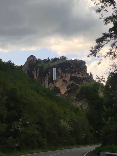 A vertical photograph shows a road curving around a mountain in Bukovo, Bulgaria, with a trans flag hanging on the mountain’s rock face.

The mountain is rocky and covered with green vegetation. A large trans flag hangs prominently from the face of the rock. The road is paved and curves through the lush green landscape. Overhanging trees and bushes obscure part of the road. The sky is overcast with heavy, dark clouds. The upper right corner shows a tree with dark, leafy branches.