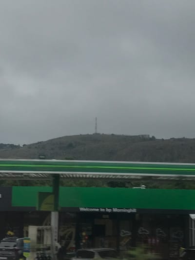 A gloomy day shows a mountain with a telecommunications tower on its peak. The sky is overcast. In the foreground, a green sign with "Welcome to bp Morninghill" printed on it is visible. A gas station, some cars, and general signs of commercial activity appear in the blurry right portion of the picture. The mountain appears bare.