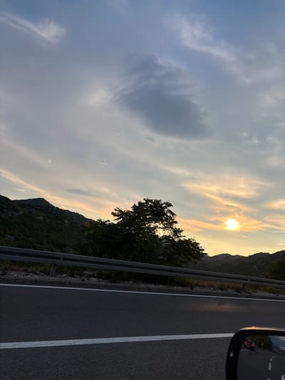 This media file depicts a view from a vehicle driving along a road in Gornja Brova, Montenegro. The scene captures a mountainous landscape bathed in the warm light of a setting sun. The sky is a mix of soft blue and wispy, golden clouds, with the sun partially obscured by them, creating a beautiful twilight effect.
On the left side of the frame, a lush, green mountainside slopes upwards, dotted with trees and vegetation. A metal guardrail runs along the edge of the road, separating it from the steep incline. On the right, the asphalt road is visible with a white line marking the lane. A side mirror of the vehicle is visible in the upper right corner, reflecting the road ahead and other vehicles.
The time of day is clearly evening, indicated by the sunset and the softer light. The weather appears to be clear with scattered clouds. The overall mood is serene and tranquil, with a sense of journey and natural beauty. There is no visible text in the image.