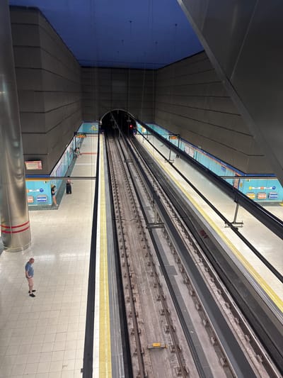 This is a high-angle, full-shot photograph of a metro station platform in San Fernando de Henares, Spain. The scene is viewed from above, looking down at the tracks and the platform. The lighting suggests it is either early morning or late evening. The platform is tiled in a light color with a yellow safety line near the edge. On the far side of the platform, there are advertisements and signage visible, including the text "SAN FERNANDO" and "LIMPIO."

Several people are visible on the platform. One person in the foreground is standing and looking towards the tracks. Further down the platform, two people are lying down, seemingly resting. Another person is sitting near the edge of the platform. The presence of people suggests the station is operational.

The train tracks are clearly visible, running parallel to the platform. Above, a slanted, dark roof structure with visible paneling covers part of the station. The overall impression is of a modern, well-maintained urban transit hub. The blue wall on the left side of the image is illuminated, creating a contrast with the darker ceiling. The perspective is slightly distorted due to the high angle of the shot.