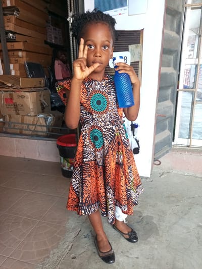 A young Black girl stands in what appears to be a storefront in Satellite Town, Nigeria. She is wearing a brightly colored African print dress, black flats, and holding a blue water bottle in her left hand. She is making a "V" sign with her right hand. Behind her are stacks of cardboard boxes and a person sitting in the background. The floor is tiled, and there is a doorway with a barred window nearby.