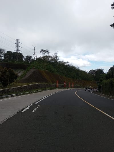 The image displays a two-lane asphalt road curving to the right, situated within the city of Phoxay, Laos. The sky is largely overcast with some visible patches of blue. The road is marked with a solid yellow center line and a solid white line delineating its left edge. A paved shoulder with a broken white line separates the main roadway from a black-and-white striped curb. Adjacent to the curb is a low, mortared stone retaining wall.

Beyond the retaining wall, a vegetated hillside rises steeply. This hillside incorporates multiple tiered earth-toned retaining wall sections, some partially covered in green growth. Five red rectangular banners with white text, spaced equidistantly, are affixed to a lower retaining wall segment. Further up the slope on the left, a set of concrete steps is partially visible, leading upwards into dense vegetation. In the upper left background, a large metal lattice electricity pylon is prominent, supporting multiple power lines, surrounded by trees and bare tree trunks.

On the curved section of the road in the middle ground, approximately three to four motorcycles, each with at least one rider, are in transit. Behind this group of motorcycles, a light-colored four-wheeled vehicle is also moving along the road. The far right side of the scene is bordered by a dense wall of green foliage.