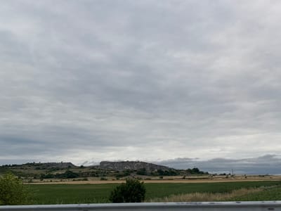 A wide shot captures a desolate, overcast landscape with a large, rocky hill formation dominating the middle ground. The hill is covered in vegetation and shows signs of human activity, possibly quarrying or ancient structures. In the foreground, a cultivated field of green crops is visible, separated by a rough, grey barrier in the lower part of the frame. The sky is filled with heavy, grey clouds, suggesting a gloomy or impending weather event. The scene appears to be a rural area, potentially near Polsko Padarevo, Bulgaria, given the rocky terrain and agricultural fields. There are no people or animals visible in the image, and the overall impression is one of natural ruggedness mixed with subtle signs of human impact.