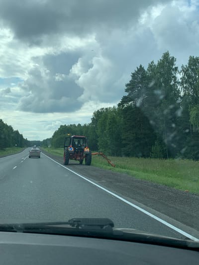 A red tractor with its door open is stopped on the side of a paved road, next to a grassy area. The tractor has a visible worker inside. A piece of orange machinery is attached to the rear of the tractor. Several cars are driving on the road, with one car in front of the tractor. The road is bordered by a dense forest on the right and trees on the left. The sky is cloudy. The location is the city of Kungur, Russia.