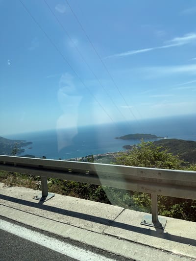 A scenic view from a vehicle, looking out over the Adriatic Sea towards islands and a coastline. The foreground features a metal guardrail, concrete barrier, and asphalt road with white markings. Beyond this, lush green hills descend to the sea, dotted with buildings and structures of a coastal town. Several small boats are visible on the water. The sky is a clear blue with thin white clouds and overhead power lines. Reflections on the window suggest the photo was taken from inside a moving vehicle. The location is identified as near Stanišići, Montenegro.