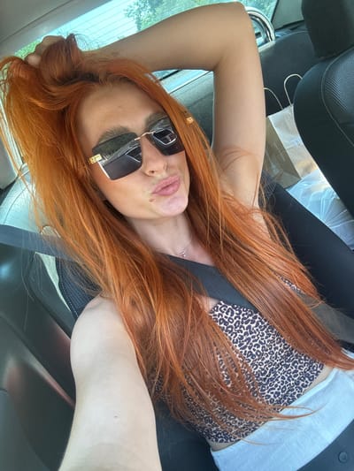 The image shows a woman with long, bright orange hair sitting in the passenger seat of a car. She is wearing large, rectangular sunglasses with a gold frame, a leopard-print top, and a light-colored garment. Her right arm is raised and resting on her head, and her left hand is not visible. She is looking at the camera and puckering her lips. A shopping bag and other items are visible in the background on the car seat. The setting appears to be outdoors, with trees visible through the car window. The woman is likely in Mišići, Montenegro, as indicated by the context.