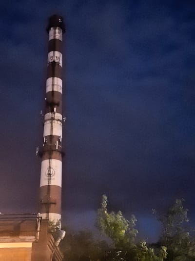 The image shows a tall, striped industrial chimney against a dark blue, cloudy sky. The chimney is predominantly white and red/brown in color, with horizontal bands. Visible markings on the chimney include the numbers "1989" and a circular logo with text that appears to read "ВЛШС". There are small structures and railings attached to the chimney at various levels, suggesting access or maintenance points. In the foreground, the roofline of a brick building is visible on the left, and trees with green foliage are present in the lower right and middle ground, some illuminated by an unseen light source. The scene appears to be taken at night or dusk. The location is identified as Novo-Talitsy, Russia.