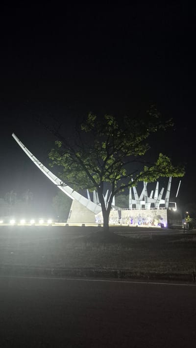 A night scene in Campo Carabobo, Venezuela, shows a modern architectural structure illuminated by lights. A tree stands in front of the structure, partially obscuring it. The area is bordered by a road and some grass. There are a few people near the structure and in an elevated section of it. The background is dark.