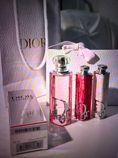 The image shows a still life arrangement of Dior products on a white surface. In the foreground, there is a box for Dior Addict perfume. To the right of the box are three Dior perfume bottles. The bottle on the far left is pink, the middle bottle is red, and the bottle on the far right is a light pink or pearl color. Behind the perfume bottles and box are two Dior shopping bags. The overall lighting is soft and diffused, with some rainbow-like reflections on the shopping bags. The scene is indoors, likely a display or collection. The location in Tomai, Moldova is not discernible from the image.