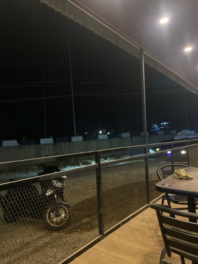 View from a covered patio area in Tanjongkapai, Indonesia, at night. A motorcycle is parked near a fence with a table and chairs on the patio. Streetlights are visible in the background.