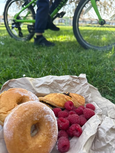 The image captures a close-up scene of a picnic in a grassy area. In the foreground, a paper bag contains two glazed donuts, a chocolate-filled pastry, and a pile of raspberries. The pastries appear to be dusted with powdered sugar. The backdrop reveals part of a bicycle, with its green frame and tire visible on the left side of the image. A person's leg is also discernible, suggesting someone is seated nearby. The background shows blurred trees and a building with a light-colored façade, indicating an outdoor urban setting, potentially in High Wycombe, United Kingdom, due to the user request. The focus is on the food, suggesting a break or picnic during a bike ride.