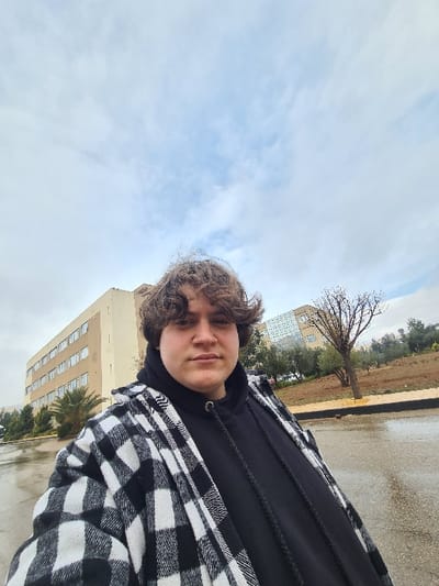 A person with curly brown hair wears a black hoodie and a black and white plaid jacket. Buildings are visible in the background, along with a bare tree and a cloudy sky. The ground appears wet. The photo seems to be taken from a slightly low angle, tilted to the side.