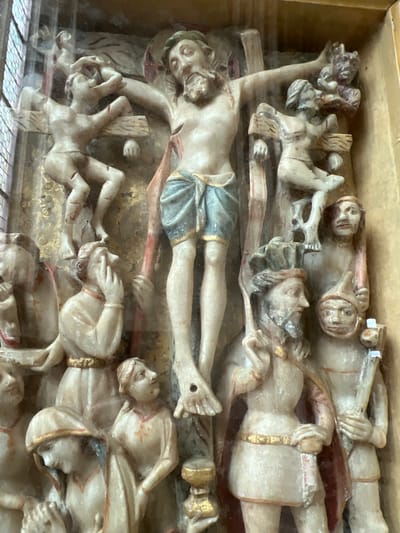 The image shows a stone carving depicting the crucifixion of Jesus Christ. Christ is shown on the central cross, with his head bowed and his body emaciated. He is wearing a blue loincloth. To his left, two figures are also depicted on crosses, their bodies contorted. One figure appears to be recoiling, while the other is reaching out. Below the crosses, a crowd of onlookers is gathered. Many of them appear to be distressed, with some covering their faces or looking up at the scene with dismay. The carving is detailed, with each figure individually rendered with distinct expressions and postures. The overall scene is one of intense suffering and communal grief. This carving is located in the Abbey of Mont Saint-Michel, France.