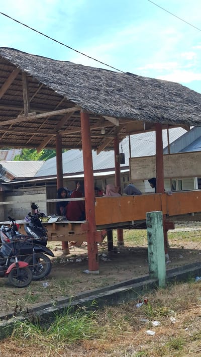 In Meunasahkrueng, Indonesia, a rustic open-air structure with a thatched roof and wooden pillars provides shade. Inside, several individuals, likely students, are seated on a wooden platform, some wearing hijabs. Some appear to be engaged in learning, with books on the platform. A whiteboard with writing is visible on the back wall of the structure. In the foreground, two motorcycles are parked. The ground around the structure is unpaved and scattered with debris. The overall scene depicts an informal educational or gathering space in a rural or semi-rural Indonesian setting, potentially part of a school or community center, with evidence of everyday life in the surrounding environment.