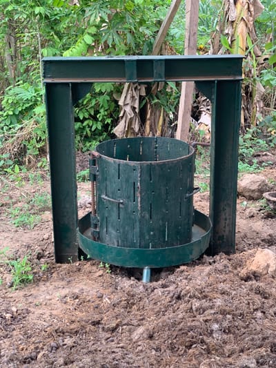 The image shows a large, green metal press, likely for extracting juice from fruit or other materials. The press is standing on a dirt ground that appears to be in a backyard or garden area. There is a dense patch of green foliage to the left of the press, possibly banana plants. The press is made of sturdy metal and appears to be in good condition. It features a round, perforated drum that rotates within a sturdy metal frame. The image appears to be taken on a sunny day, as there is no shadow cast by the press. The coordinates 7.119, 5.362 are located in the Egbeda area of Lagos, Nigeria.