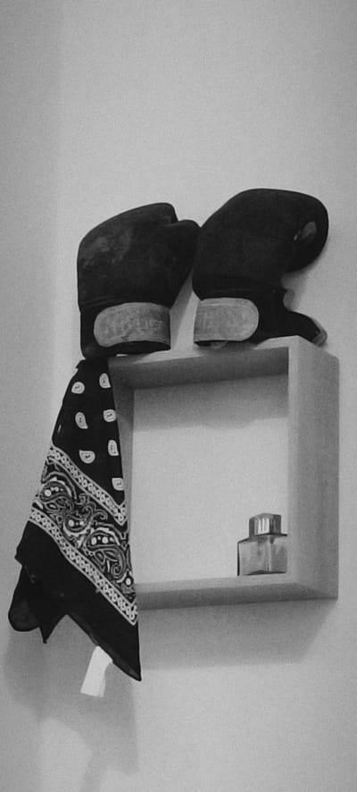 A pair of black boxing gloves rests on the top edge of a white, recessed shelf mounted on a white wall. A black bandana with a white paisley pattern hangs from the left glove, draping down in front of the shelf. On the right side of the shelf, a small, clear glass bottle with a silver lid is visible. The lighting is dim, and the image is grainy. The scene is devoid of people and activity, suggesting a still life arrangement or an item left behind. The location is the city of Winnenden, Germany.