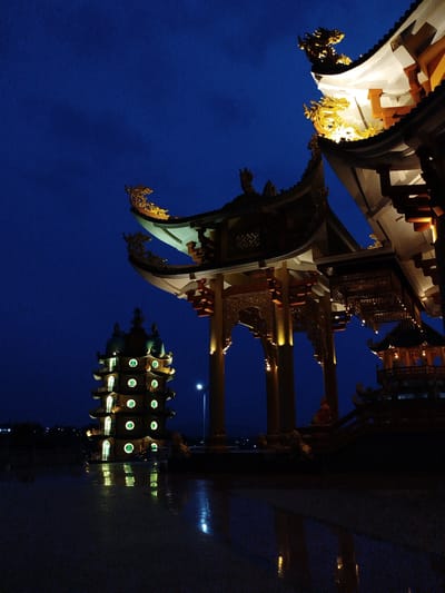 The image depicts a nighttime scene in Thiện Chí, Vietnam, showcasing illuminated Chinese-style architecture. A pagoda with multiple tiers and circular windows emitting a green light is on the left. A more elaborate structure with ornate, golden dragon figures on the roof edges is partially visible on the right, its roof illuminated with bright light. The ground appears to be wet, reflecting the lights from the buildings. The sky is a deep blue. There are no people visible in the scene.