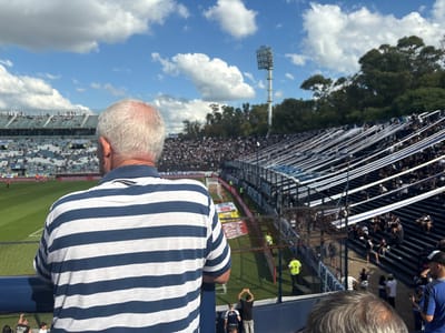 The image captures a back view of a man with short gray hair, wearing a navy blue and white striped polo shirt, leaning on a dark railing. He is looking out onto a bustling football stadium filled with spectators. The perspective is from an elevated section of the stands, offering a wide view of the event.
The stadium features multiple tiers of seating, mostly dark blue or black, packed with a large crowd of people. Numerous white and blue banners or streamers are draped vertically along the railings of the stands, suggesting team colors or decorations for the event. In the distance, on the left side, another section of the stadium is visible, also filled with fans.
The field, a vibrant green, is partially visible at the bottom and to the left, with goalposts and markings indicating a football (soccer) pitch. A few individuals in bright yellow safety vests can be seen near the field perimeter. Above the stadium, the sky is bright blue with scattered, fluffy white cumulus clouds, indicating a clear and sunny day, likely mid-day or early afternoon. A tall stadium light tower stands prominently against the sky on the right. Lush green trees line the distant edge of the stadium.
A red advertising banner near the field prominently displays the word "MOSTAZA." The overall scene depicts a lively and well-attended outdoor sporting event.