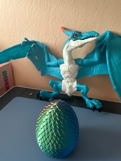 A close-up, eye-level shot shows a blue and white toy pterodactyl with its wings spread, positioned above a textured, iridescent dragon egg. The toy pterodactyl has a white chest and neck, with blue limbs and wings. Its head is blue with black eyes and a prominent crest tipped with orange. The dragon egg is spherical and covered in a scaled pattern, reflecting hues of green and blue. The objects are placed on a flat, dark surface, possibly a tablet or tray, which rests in front of a beige wall with a textured finish. The lighting suggests an indoor environment, with the wall illuminated from the left. There are no people or animals visible in the image. The scene does not provide any geographical context or information that would allow for its placement in Hradec Králové, Czechia.