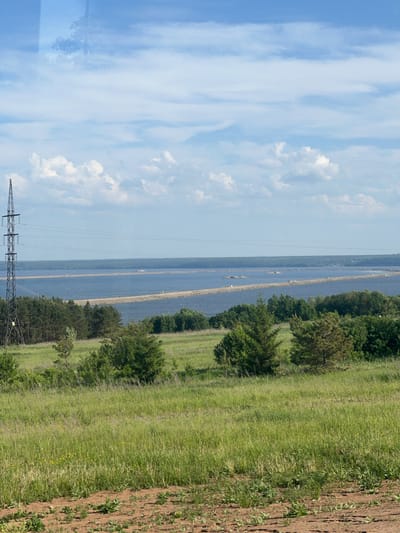 The scene is a rural landscape in Белоус, Russia, featuring a river with two narrow, sandy islands running parallel within it. A high-voltage transmission tower is visible in the lower left corner of the frame. To the right of the river are patches of trees and green grassy fields, extending to a section of tilled soil or a dirt road. The sky occupies the left side of the image and is blue with streaks of white clouds.