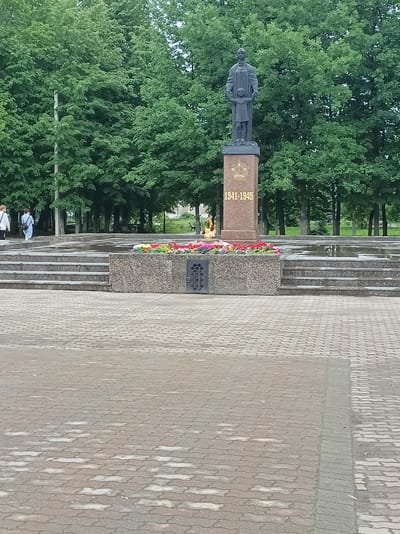 This is a full, eye-level, outdoor, daytime, vertical, medium shot of a war memorial in Buy, Russia. In the background, there is a dense forest of green trees. In the center of the frame, a tall, dark-colored statue of a woman holding a child stands on a reddish-brown, square pedestal. The pedestal has a gold star and the dates "1941-1945" inscribed on it. In front of the pedestal is a low, granite wall adorned with a colorful flower arrangement. To the left of the memorial, there are tiered, concrete steps leading up to a water feature. Two individuals, one wearing a white shirt and the other a blue dress, are walking up the steps. The foreground consists of a paved plaza with interlocking gray and reddish-brown bricks. The scene is tranquil and solemn, evoking a sense of remembrance for fallen soldiers.