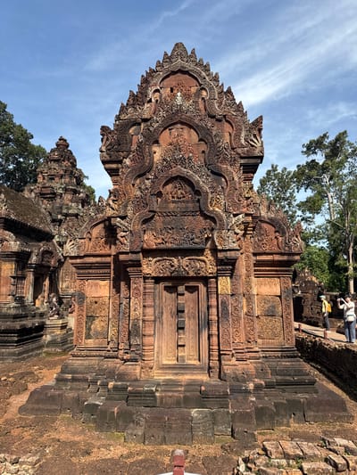 The image shows the central sanctuary of Banteay Srei temple in Siem Reap, Cambodia, a 10th-century Hindu temple dedicated to Shiva. The structure is made of red sandstone and features intricate carvings. The central doorway is prominently displayed, framed by ornate carvings of deities and mythical creatures. The surrounding grounds are covered in red earth and scattered stones, with trees visible in the background under a blue sky with scattered clouds. Two individuals, a woman wearing a yellow backpack and a person with a camera, are standing on a wooden walkway to the right of the main structure, observing the temple.