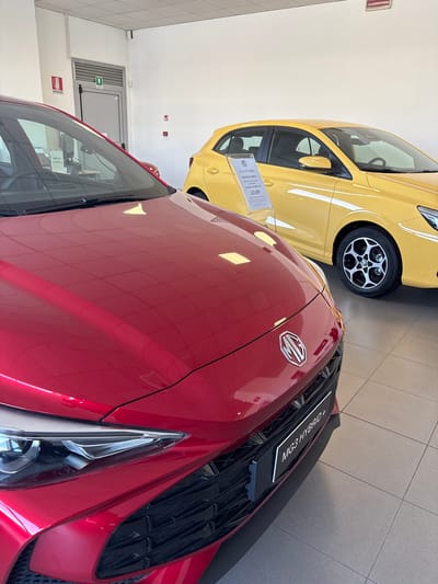 This media file captures an indoor view of a car showroom in Ancona, Italy, featuring two compact cars on display. The foreground is dominated by the front portion of a red metallic car, likely an MG, showcasing part of its hood, grille, and a headlight. A sign positioned on the grille clearly reads "MG3 HYBRID +". The car's polished surface reflects the indoor lighting.

Behind the red car, a yellow hatchback is partially visible. This car also appears to be an MG, as indicated by the logo on its wheel. A promotional sign stands beside the yellow car, displaying information such as "GRUPPODIBA", "PROMO MG3 BENZINA", "47 RATE A PARTIRE DA", and a price of "133,00€".

The setting features light-colored tiled flooring and white walls. In the background, an emergency exit door with a green "EXIT" sign is visible, along with a red "ESTINTORE" (Italian for fire extinguisher) sign mounted on the wall. The bright indoor lighting suggests the cars are presented for sale or viewing in a dealership. No people are visible in the image.