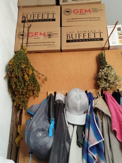 The media file depicts a close-up, vertical shot of a wall with several objects hanging on it. In the background, there are stacked cardboard boxes, some with the logos of "THE HOWARD G. BUFFETT FOUNDATION" and "GEM GLOBAL EMPOWERMENT MISSION UKRAINE." The boxes appear to be storage or shipping containers.
Hanging from wooden pegs are several items. On the left, a large bunch of dried yellow flowers and greenery, possibly herbs, hangs prominently. To its right, a grey drawstring bag with blue accents is suspended. Further to the right, a white baseball cap with a mesh design is visible, alongside various folded fabrics and towels in blue, pink, and beige tones. A small bunch of dried white flowers is also hanging on the right side of the frame.
Visible text includes "THE HOWARD G. BUFFETT FOUNDATION," "GEM GLOBAL EMPOWERMENT MISSION UKRAINE," "www.globalempowermentmission.org," and "#GemMissions." There are also handwritten annotations in what appears to be Cyrillic script, such as "круп сахар" (likely "coarse sugar").
The scene suggests an indoor setting, possibly a storage area or a home, given the variety of objects. The lighting is diffused, indicating it is likely daytime but without direct sunlight. The overall impression is one of organized storage with decorative elements of dried plants. The mention of Ukraine in the text suggests the location context.
