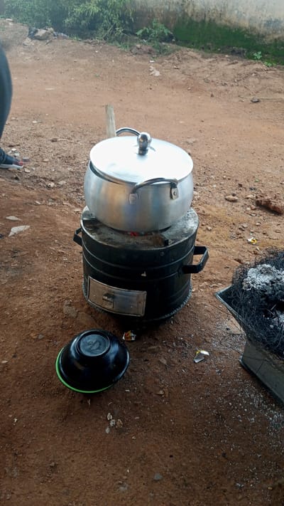 An outdoor scene in Barikin Sabon Gidan Kanat, Nigeria, depicts a cooking area established on reddish-brown dirt ground. A silver-colored metal cooking pot with a lid is positioned on a black, cylindrical metal stove. The stove features side handles and a rectangular access door on its lower front; glowing red embers are visible beneath the pot, indicating active combustion. To the left and foreground of the stove, an inverted black plastic bowl with a visible green rim rests on the ground. To the stove's right, a square metal container holds dark, burnt debris topped with white ash. The ground surface is irregular, composed of dirt, small stones, and scattered white fragments. In the upper-left, a portion of a human foot, wearing a dark shoe with a white sole and red-painted toenails, is partially visible. The background reveals a light-colored, possibly stuccoed wall, behind which green vegetation grows.