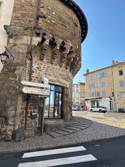 The image depicts a street scene in Le Puy-en-Velay, France, characterized by historical architecture and urban infrastructure under a clear blue sky.
In the foreground, a white pedestrian crossing with two thick stripes spans an asphalt road. Adjacent to this, on the left, is a prominent, multi-story stone building with a rounded, fortified-style corner section. The lower facade consists of rough-hewn stone blocks, transitioning to more regular ashlar masonry on the upper levels. A black, decorative street lantern is affixed to its left-hand wall, and a grey downspout runs vertically along the building's corner. Below a decorative stone architrave, a modern entrance features a dark-framed glass door. To the left of this door, a brown metal pole holds two white directional signs. The top sign, pointing left, reads "Eglise du Collège." The bottom sign, pointing right, indicates "Marché Couvert" and "BIBLIOTHEQUE." A black wall-mounted mailbox is positioned below these signs. Curved stone steps lead from the cobblestone sidewalk up to the building's entrance. Above the doorway, the rounded building section is supported by a series of intricately carved stone corbels, topped by a section of lighter stone blocks and a small, rectangular window with a red-brown frame.
The cobblestone street curves to the right, leading towards an intersection. A white SUV is parked beyond the pedestrian crossing on the right side of the street, facing away from the viewer. A black traffic light pole stands on the right sidewalk. In the background, several traditional multi-story European buildings are visible, including a cream-colored structure with light blue shutters on its windows and a light yellow building with similar architectural features and red-tiled rooftops. No individuals are present in the scene. The ambient light suggests daytime conditions.