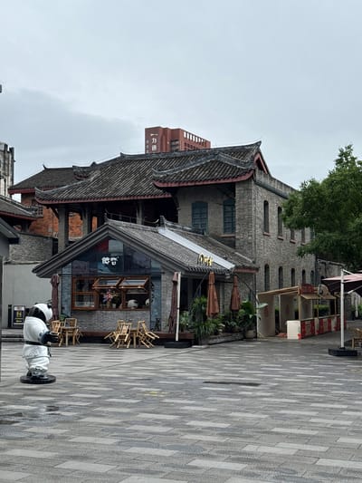 This image depicts an outdoor urban environment within Qingyang District, China, under an overcast sky. The foreground consists of a large plaza paved with rectangular grey and dark grey interlocking stones, which show visible damp patches.

On the left side of the plaza, a life-sized statue of a panda is positioned, dressed in a white traditional Chinese martial arts robe with blue trim and a black sash, standing on a black circular base. Behind the panda, a blue sign with white pictograms for restrooms (male, female, and an arrow) is attached to a pole.

In the midground, a single-story commercial building with light grey brick walls and a grey tiled gabled roof is prominent. Its facade is largely glass, displaying the white Chinese characters "德睿" (Dé Ruì). Several wooden-framed windows, some open, reveal interior decorations including flowers and a small panda figure. Outside this building, multiple wooden tables and chairs are arranged on the pavement, some stacked, along with folded brown outdoor umbrellas. Above the entrance of this structure, gold-colored Chinese characters are visible on the roof's edge.

Behind this commercial structure, a larger, multi-story building with a darker grey tiled roof, pronounced eaves, and reddish-brown architectural accents is partially visible. Its walls are constructed of dark brick. To the right, another multi-story building with dark grey brick walls and a matching tiled roof features a series of tall, narrow arched windows with dark frames. A large, dense green tree partially obstructs the view of this building on the far right.

Further to the right on the plaza, several temporary outdoor vendor stalls are erected, characterized by light-colored canopies and red and white patterned skirting, all appearing vacant. In the distant background, a modern, reddish-brown high-rise building is visible, bearing the vertical white Chinese characters "力迅" (Lì Xùn) on its facade. The entire scene is devoid of any visible human presence or activity.