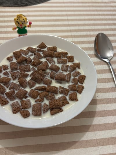 A close-up shot shows a white bowl of chocolate cereal in milk, positioned on a table with a striped cloth. To the left of the bowl, a small, yellow, tiger-like figurine with a green dress and a pink flower accessory is visible. To the right of the bowl, a silver spoon rests on the cloth. The background is blurred, with a dark gray textured surface at the top. The scene is set in a domestic environment within the city of Banići, Croatia.