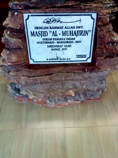 A white marble plaque with black lettering, mounted on a rough, brown, rock-like structure, sits on a light brown tiled floor. The plaque reads "DENGAN RAHMAT ALLAH SWT. MASJID ''AL - MUHAJIRIN'' PERUM RENDOLE INDAH MUKTHIHARJO - MARGOREJO - PATI DIRESMIKAN OLEH BUPATI PATI H. HARYANTO SH.MM.M.Si. 03 SYAWAL 1440 H 12 OKTOBER 2019". The plaque is likely located in a mosque at the address -6.738, 111.014 in Kecamatan Margorejo, Indonesia. The plaque likely commemorates the opening of the mosque.
