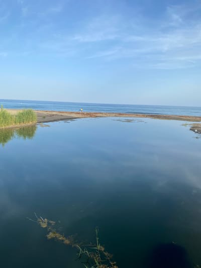 The image depicts a tranquil scene at the Resinera-Voladilla, Spain. A narrow strip of sandy shore separates a calm, deep blue body of water from a clear sky dotted with wispy white clouds. The water reflects the sky, creating a mirror-like effect.
On the shore, a small figure in a yellow top is visible, appearing to be a person. Further along the shore, green vegetation grows, its reflection also visible in the water. In the deeper water, some floating debris and aquatic plants can be seen. The lighting suggests it is daytime, possibly late afternoon, given the soft light and the clear, bright sky. The overall mood is peaceful and serene, with a sense of stillness and natural beauty. There is no visible text in the image.