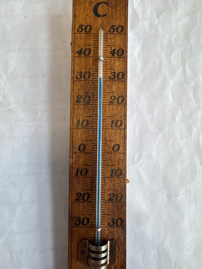 The image shows a close-up of a wooden thermometer. The thermometer is marked with Celsius degrees, ranging from 10 to 50. The blue liquid inside the thermometer is visible, indicating the current temperature. The thermometer is positioned vertically, and there are some faint markings on the paper behind it, which appear to be printed text, possibly from a document or book. The thermometer appears to be an old-fashioned, analog device. The specific location in the city of Mejaniga, Italy, is not discernible from the image itself.
