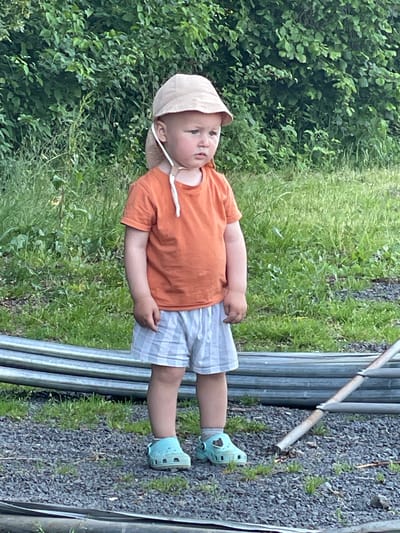 The image shows a young child, likely a toddler, standing outdoors. The child is wearing a light-colored sun hat, an orange short-sleeved t-shirt, and checkered shorts. They are also wearing light blue Crocs. The child appears to be looking off to the side with a neutral or slightly curious expression.
The setting is natural and somewhat rustic. Behind the child, there is lush green foliage, possibly bushes or trees, and tall green grass. In front of the child and to their right, there is a gravelly ground surface and a series of parallel metal pipes or railings. The lighting suggests it is daytime, possibly late afternoon or early evening, given the quality of light. The overall atmosphere seems peaceful and casual, suggesting a child playing or exploring outdoors. No text is visible in the image. The location context is Beltershain, Germany.