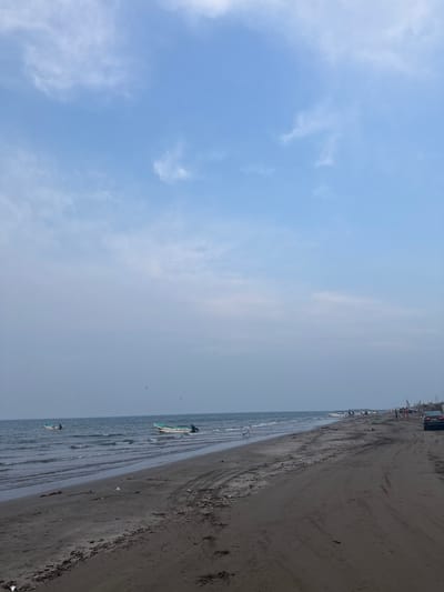 The image displays a wide, ground-level view of a beach in Antón Lizardo, Mexico, under a light blue sky with scattered, wispy white clouds. The ocean, characterized by blue-grey water and small waves, occupies the left side of the frame. Two small, white boats with light green trim are positioned in the shallow water near the shore in the mid-ground, one featuring a visible outboard motor. The beach itself is composed of dark grey sand and dirt, extending from the foreground into the distance on the right, and exhibits multiple vehicular tire tracks. Scattered debris is present along the waterline and on the sand. In the far distance on the right, a dark-colored sedan is parked on the sandy path, and several indistinct human figures are visible on the beach. Beyond these figures, additional small boats are either in the water or pulled onto the sand, adjacent to what appear to be rudimentary structures constructed from wood and tarpaulins. A single piece of metallic debris is partially submerged near the water's edge in the mid-ground.