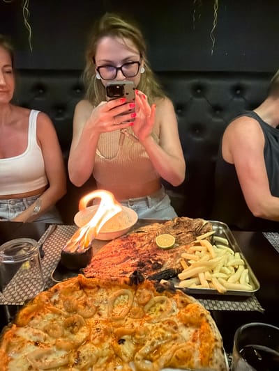 The image captures a dining scene in Maafushi, Maldives. Three individuals are seated around a table covered in a textured surface. A woman with blonde hair and glasses is holding up a smartphone, presumably to take a photo of the meal. She has bright red nail polish. A second woman, with similar blonde hair, is wearing a white tank top and denim shorts, while the third person is wearing a gray tank top. A large platter dominates the table containing a grilled fish topped with a lemon wedge, a side of fries, and a pizza with banana slices. A decorative flame holder, containing an open flame, sits between the fish and the pizza. Glasses are also visible on the table. The background features a cushioned wall.