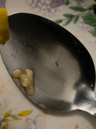A close-up shot reveals a slightly used metal spoon resting on a patterned plate. A small amount of pale, yellowish food residue clings to the spoon's surface, possibly leftover from a meal. The plate displays floral designs in soft hues of green, purple, and yellow. The spoon's reflective surface shows minor scratches and imperfections, suggesting it has been used multiple times. A portion of a yellow object is present in the bottom left corner.