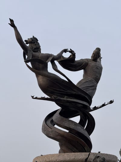 The image depicts a bronze sculpture, specifically a component of the "Long Mark" sculpture located at the Lintong Museum in the Lintong District, China. The sculpture features a standing figure in traditional Chinese attire grasping the hand of another figure below, who is reaching downwards. The figures are bronze and stand against a light sky. The sculpture also incorporates swirling, metallic ribbons with decorative patterns. To the right, a partially visible cylindrical column with carved embellishments is present.