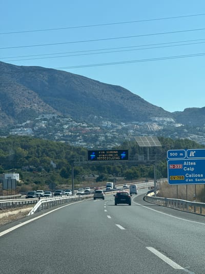The image shows a view from a car on a highway in Altea, Spain, on a sunny day with a clear blue sky. The road has multiple lanes, indicated by white dashed lines. In the distance, a town with white buildings is built on a hillside, covered by a large mountain. There are trees on the lower slopes of the mountain.
A large electronic road sign displays "FIN SEMANA" (end of week) in Spanish and "VELOCIDAD MOTO CICLISMO" (motorcycling speed) with blue arrows and speed indicators. To the right of the electronic sign, a blue road sign indicates directions to "Altea," "Calp," "Callosa," and "d'en Sarrià" via highways N-332 and CV-755. Another sign indicates "500 m" with an arrow. A solar panel array is visible behind the electronic sign.
Several vehicles are on the highway, including cars and trucks. The overall environment is a highway leading towards a Mediterranean coastal town.