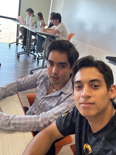 A photograph depicts two young adult males in the foreground of what appears to be a modern educational or collaborative workspace in Fraccionamiento Los Heroes Queretaro, Mexico. The male on the left, with dark hair and a light complexion, wears a blue, red, and white plaid long-sleeved shirt with a black choker necklace featuring a small gold cross. He is seated at a light-colored desk with an orange chair and looks slightly to his right. The male on the right, also with dark hair and a light complexion, wears a black short-sleeved shirt with a gold "Venum" logo on the chest. He is positioned closer to the camera, appearing to capture the image, and looks directly forward with a slight smile.
In the background, four additional individuals are seated at a long, rectangular glass-top table with metal, wheeled bases and orange chairs. From left to right at this table: a male in a green shirt is looking at a pink laptop next to a white tumbler; a female in a light-colored top is looking towards the right, with a clear water bottle on the table; a person in a dark top is partially obscured; and another male in a white shirt with white earbuds is looking towards the left.
The environment features a light brown tiled floor, a plain white wall on the right, and large glass windows on the left that reveal an outdoor area with green vegetation under bright daylight. The overall scene suggests an active study or work session.