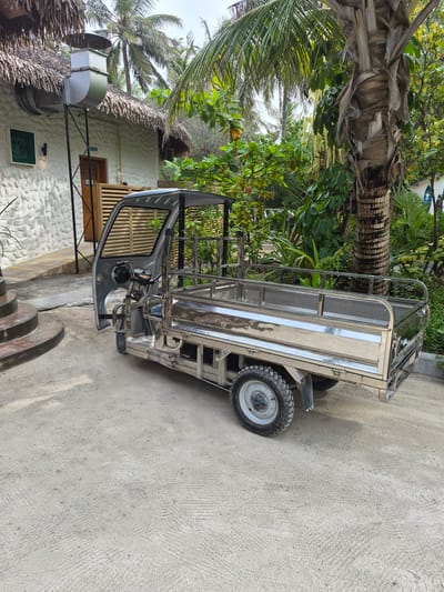 The image shows a three-wheeled vehicle, likely a utility vehicle or small truck, parked on a concrete surface in Huraa, Maldives. The vehicle has a metal frame and a small cargo bed. It is positioned near a building with a thatched roof, indicating a tropical environment. Palm trees and other greenery are visible in the background. The scene appears to be outdoors in a residential or commercial area. The image is taken at an angle, likely by a pedestrian.
