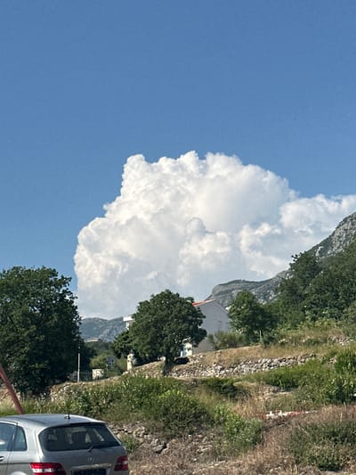This is a vertically oriented, outdoor, daytime photograph taken in Blizikuće, Montenegro. The scene depicts a large, fluffy white cumulus cloud against a bright blue sky. To the right of the cloud, a steep, green mountainside is visible, with trees and rocky terrain. A stone wall runs along the lower part of the mountainside. In the foreground, the rear of a silver minivan is visible, parked on a dirt or gravel area. A red railing is also partially visible in the bottom left corner. The overall impression is a scenic, natural landscape under clear, sunny weather. There are no people clearly visible in the image, and no discernible text.