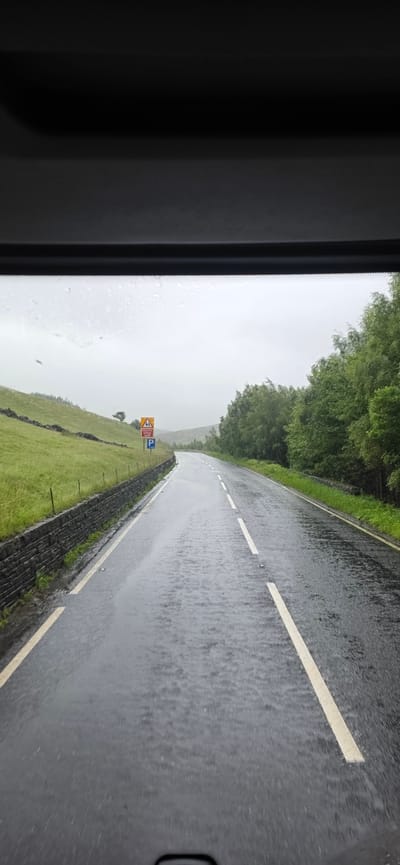 The image shows a wet, paved road in the city of High Peak, United Kingdom, during what appears to be rainy or overcast weather. The road surface is dark and shiny, reflecting the grey sky and the surrounding environment, with visible streams of water running along it. The road has white dashed lines on the left and right edges and a single white dashed line down the center, indicating it is a two-lane road.
To the left of the road, there is a steep, grassy embankment with a low stone wall running along its base, separating it from the road. Further up the embankment, there are scattered trees. On the embankment, near the road, there are two signs. The upper sign is triangular with a yellow background and a black symbol depicting a series of curves, indicating a winding road ahead. Below this sign is a blue rectangular sign with a white 'P', indicating parking is available.
To the right of the road, there is a dense line of lush green trees, forming a backdrop to the scene. The sky is overcast and grey, contributing to the damp atmosphere. The overall impression is of a rural or semi-rural setting in High Peak, captured from the perspective of someone inside a vehicle, looking forward.