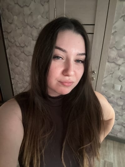 The image is a close-up, vertically oriented selfie of a young woman with long, dark brown hair. She is wearing a dark brown, high-necked top. Her face is turned towards the camera, with her head tilted. She has fair skin, dark eyebrows, and blue eyes. Her lips are slightly parted, and she appears to be looking directly at the camera with a neutral or slightly playful expression.
The background consists of a wall with floral wallpaper in muted tones of grey and white. To the left of the woman, a closet door or cabinet is visible, featuring a light grey finish with two silver knobs. Above the closet, there is a horizontal trim or shelf. The lighting in the image suggests it was taken indoors, possibly during the daytime or with artificial lighting. There is no visible text in the image. The provided location context is Гамово, Russia.