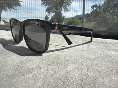 A pair of black Gucci sunglasses with gold branding and a red and green accent on the left temple are placed on a light gray, textured surface, possibly a car dashboard, with the lenses reflecting the sky and trees. In the background, a wire fence is visible, beyond which are trees and hills under a partly cloudy sky. The scene is outdoors, with sunlight casting a shadow from the sunglasses. The location is stated as the city of Tekeli, Kazakhstan.